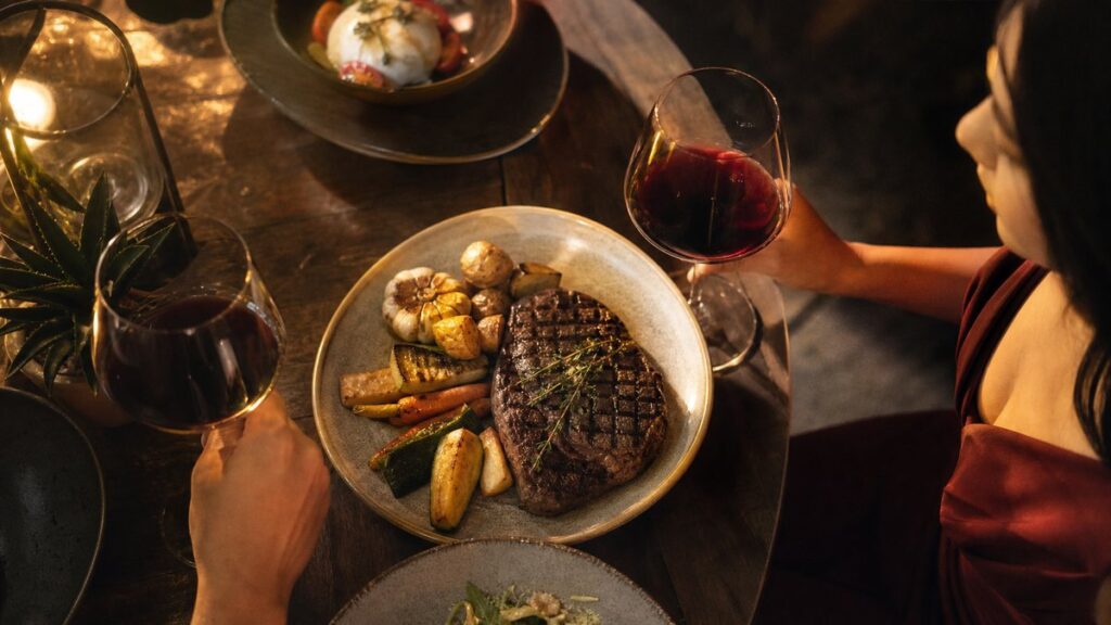 Overhead view of a restaurant table where a grilled steak, roasted vegetables, and two wine glasses are arranged in a balanced, natural spread. Warm evening light falls across the plate, while deep burgundy and subtle gold tones in the food and wine suggest richness, harmony, and a thoughtfully paired meal.