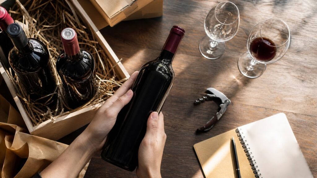 An overhead process scene on a wooden tasting table where unopened wine bottles, shipping paperwork, and a corkscrew are arranged around a few clean glasses. Natural window light highlights the bottle glass and liquid tones, creating a calm, analytical mood that hints at quality preserved through smart sourcing, not compromise.